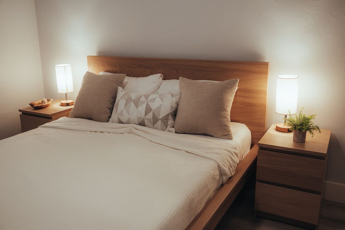 A quiet bedroom at dusk with a neatly made bed and a softly glowing bedside lamp